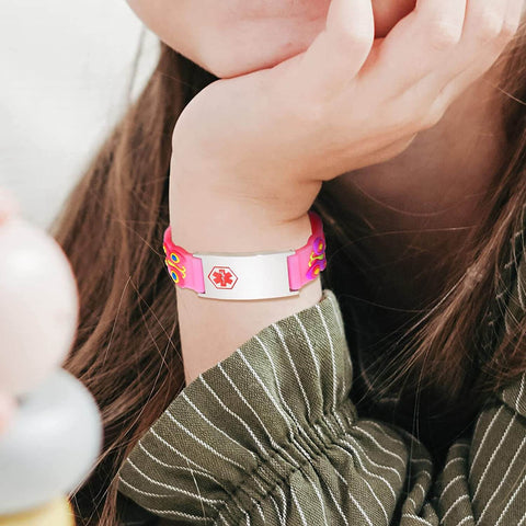 Child wearing child medical alert bracelet. Bracelet band is made of pink silicone with a butterfly design. The alert plate is a stainless steel with red medi-alert symbol engraved on the plate.